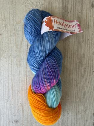 Ein handgefärbter Garnstrang in kräftigem Blau mit Farbverläufen in Orange, Türkis und Pink liegt auf einem hellen Holzuntergrund. Das Garn ist mit einer Herzfasern-Banderole versehen.